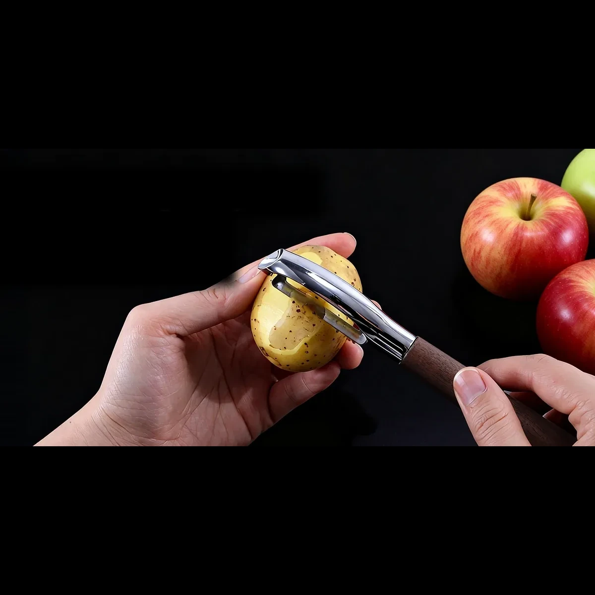 Y-shape wood-handle peeler removing skin from a potato with apples on a dark prep surface